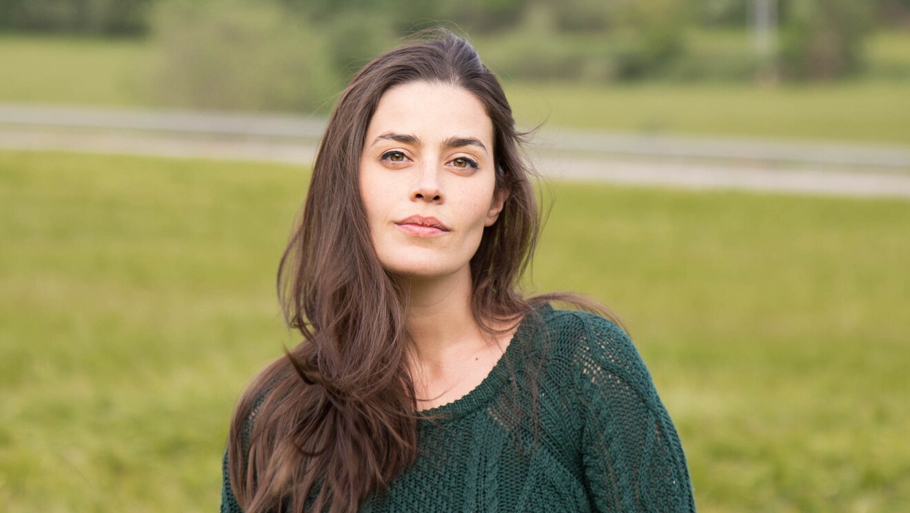 Lorena Franco es escritora y empezó autopublicándose en 2016 con el servicio KDP de Amazon. En un parque con fondo de árboles y césped está un plano corto de Lorena con el pelo castaño y muy largo y al viento. Tiene los ojos marrones azules y mira a cámara sin sonreir. Lleva un jersey de punto calado de color verde. 