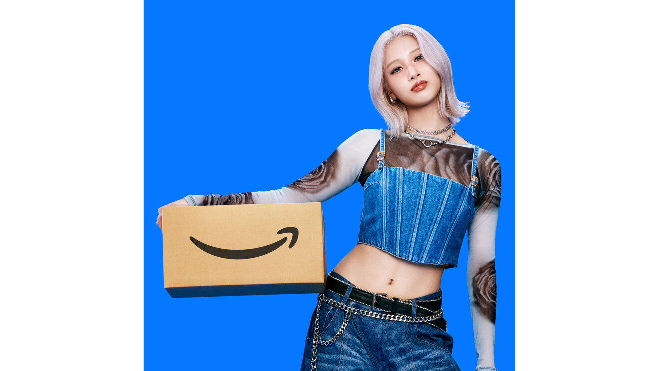 ハイトーンカラーヘアの若い女性が、Amazonロゴ入りダンボール箱を右手と腰で挟んで持っている。