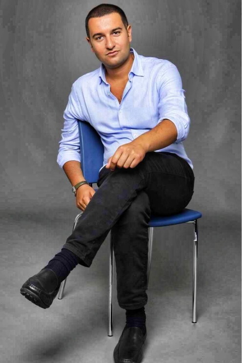 Un uomo seduto su una sedia. Indossa una camicia celeste e dei pantaloni scuri. Lo sfondo della foto è grigio.