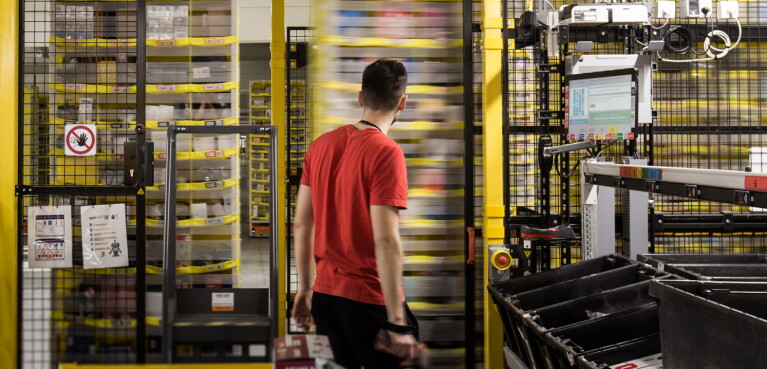 Un dipendente Amazon ripreso di spalle al centro dell'immagine osserva gli scaffali mobili del magazzino. 