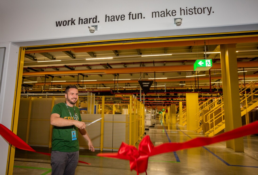 Ein großes, geöffnetes Eingangstor zeigt den Blick in eine Lagerhalle: Man sieht gelbe Treppen und einen gelb eingezäunten Regalbereich. Links im Bild ist der Standortleiter zu sehen. Er trägt ein grünes T-Shirt mit Aufschrift "FRA7" und hält eine Schere in der Hand. Er ist im Begriff, ein riesiges rotes Schleibenband, das am Eingangstor angebracht ist, zu zerschneiden.