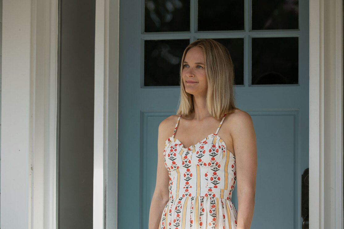 Susannah from "The Summer I Turned Pretty" stands on her porch in a white sundress with colorful patterns looking at something off camera.