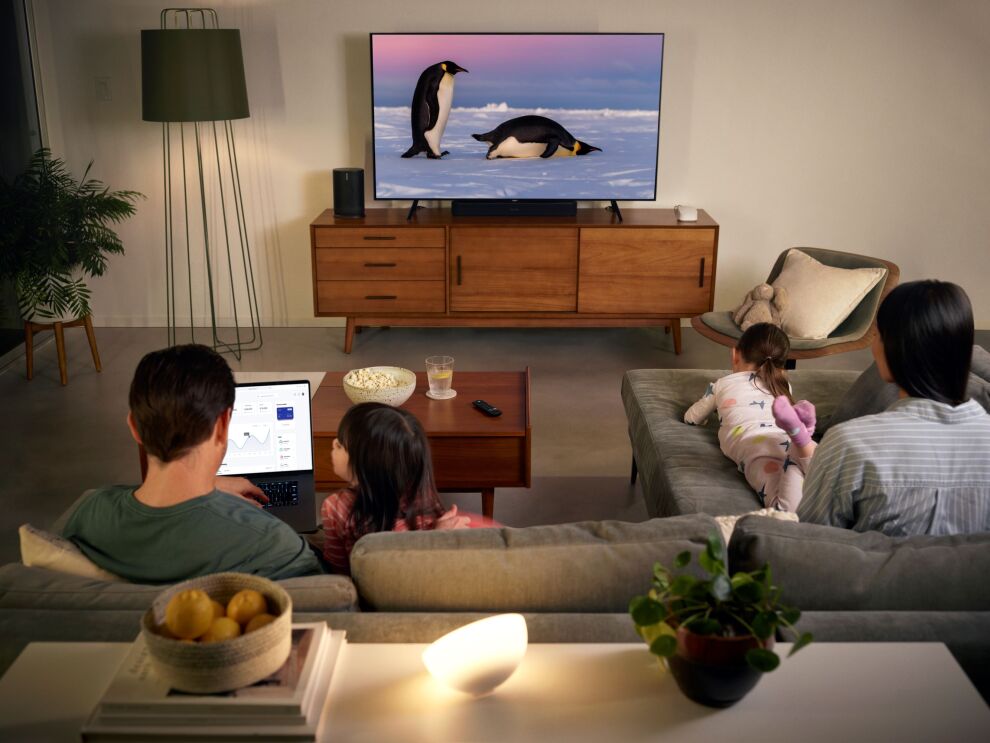 リビングルームに4人家族がテレビの前に集まっている。みんなはロングL字型のソファに座っている。テレビにペンギンの画像が表示されている。