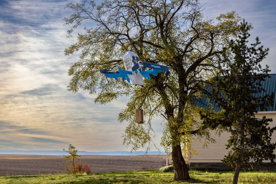 A Prime Air drone carries an Amazon parcel in the air in front of a tree and cloudy sky