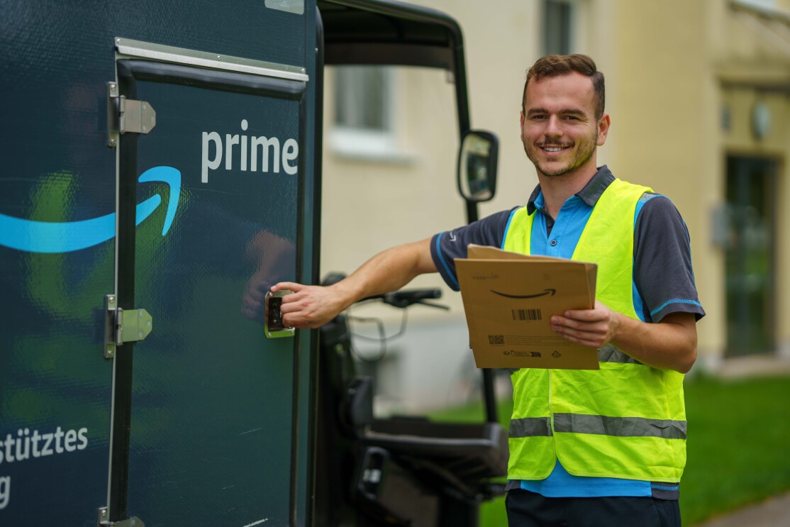 Ein junger Amazon-Fahrer mit dem E-Cargo Bike: Er trägt Berufskleidung im Amazon-look und ist mit einem blau-grauen E-Lastenrad unterwegs.