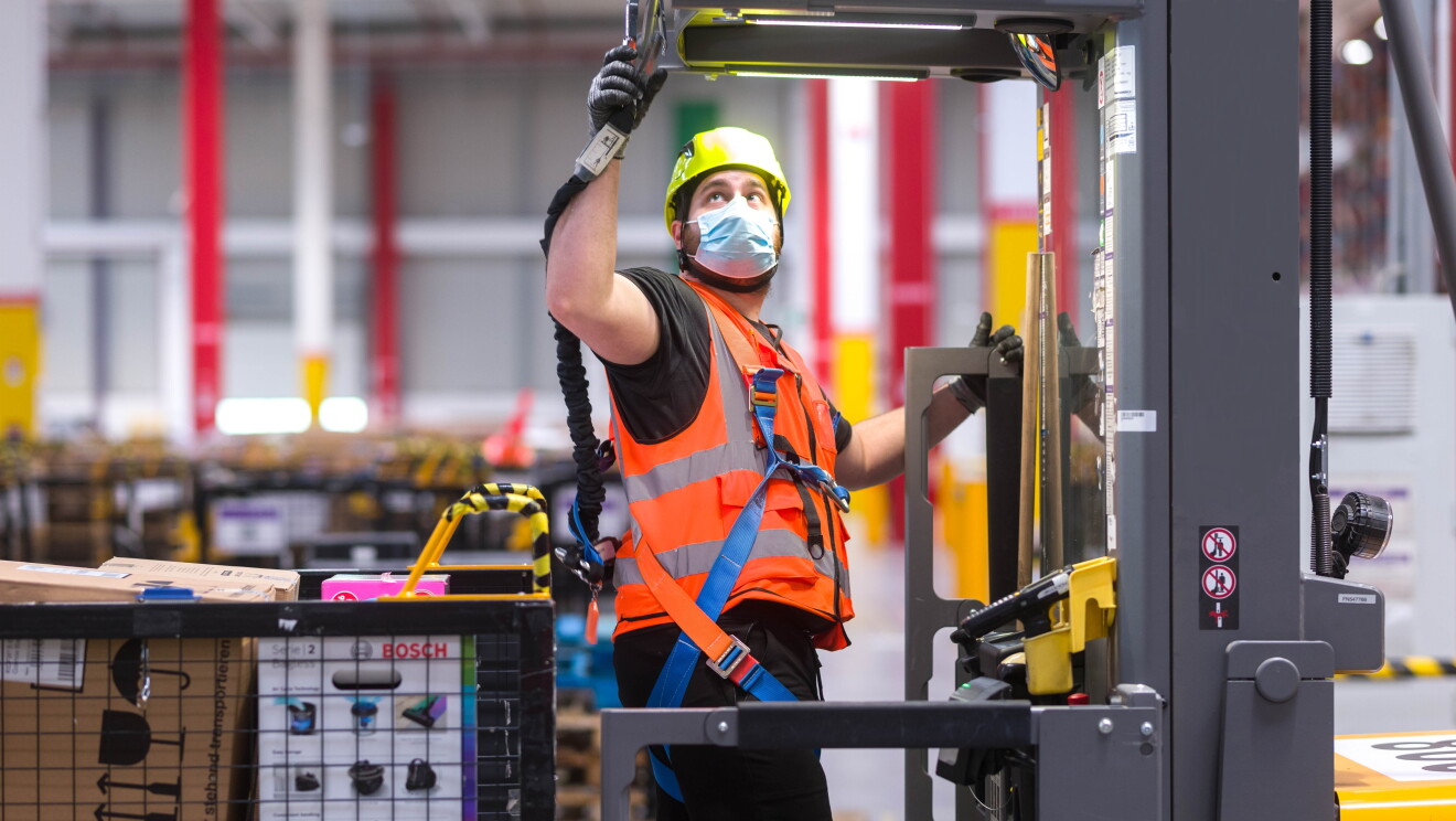 Dipendente di un magazzino Amazon. Indossa un casco, una mascherina chirurgica e un giubbino di sicurezza, si trova al centro di un macchinario. Alle sue spalle un cesto con dei pacchi Amazon. 