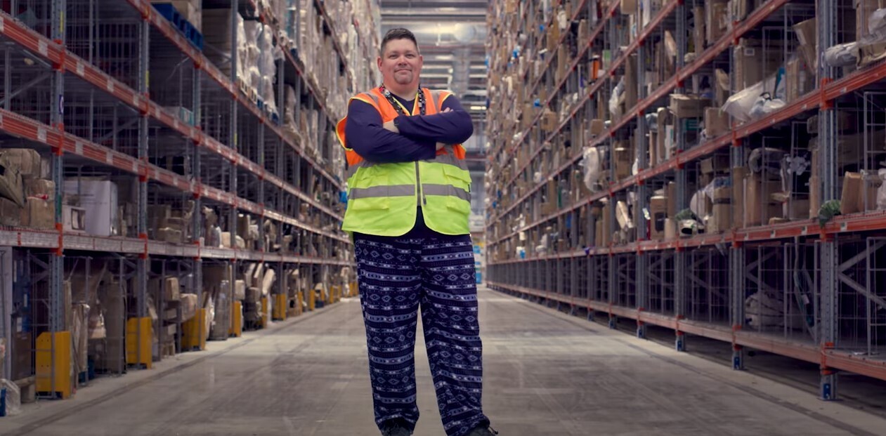 Burnell wears a safety vest and pajama pants in an Amazon fulfillment center. He crosses his arms and smiles.