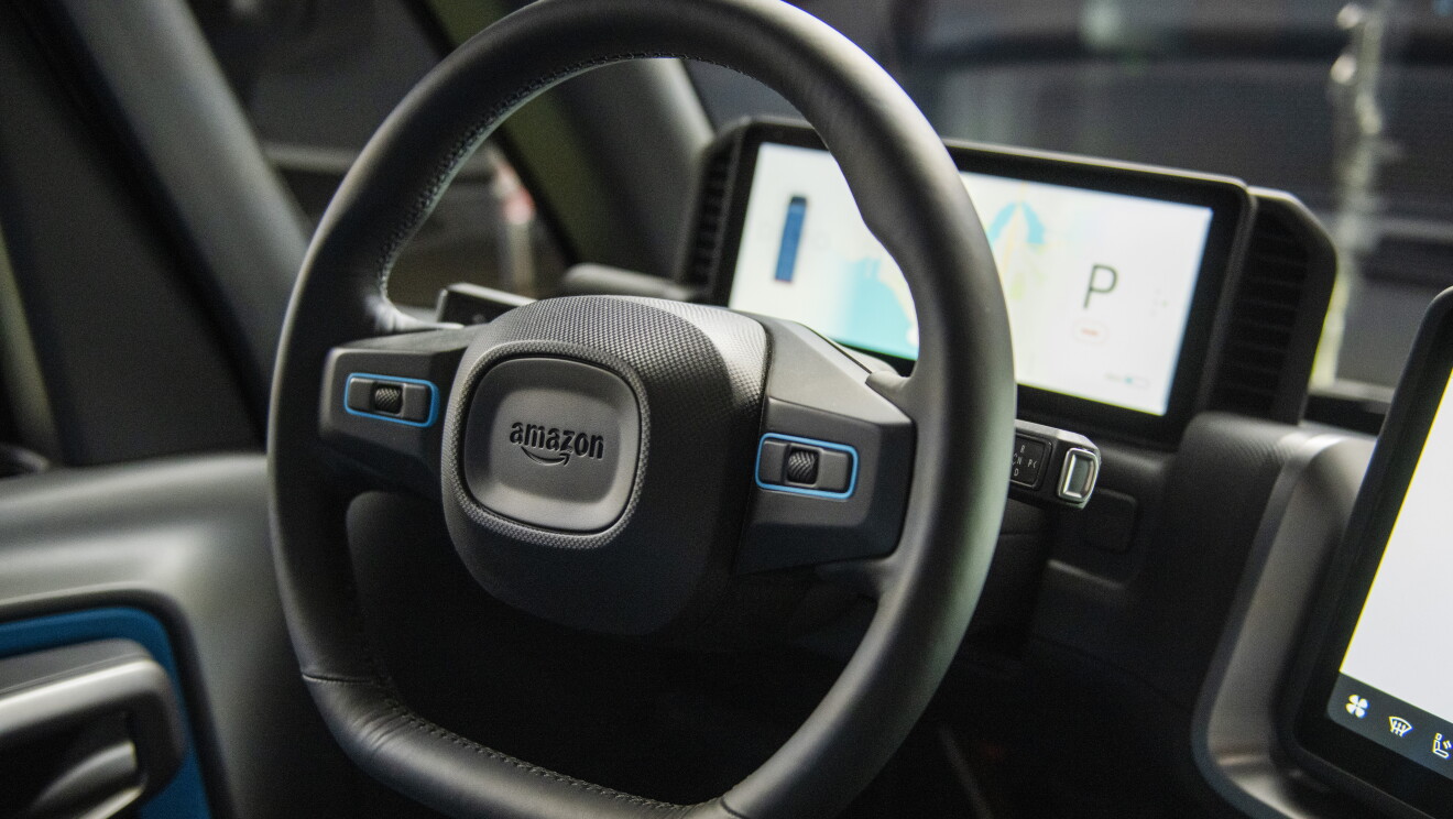 Primer plano del volante de la furgoneta Rivian. En el centro del volante está el ligi de Amazon en el fondo una pantalla de navegación. El volante es de color negro. 