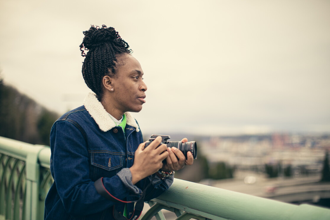 A woman in a blue jacket and green top, and holding a camera, looks to the right from atop a bridge. 
