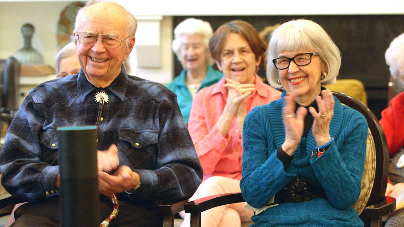Two retirees, a woman in a blue dress and a man in a blue and black flannel short and bolero, laugh and clap at a performance. A black Amazon Echo is in the foreground.