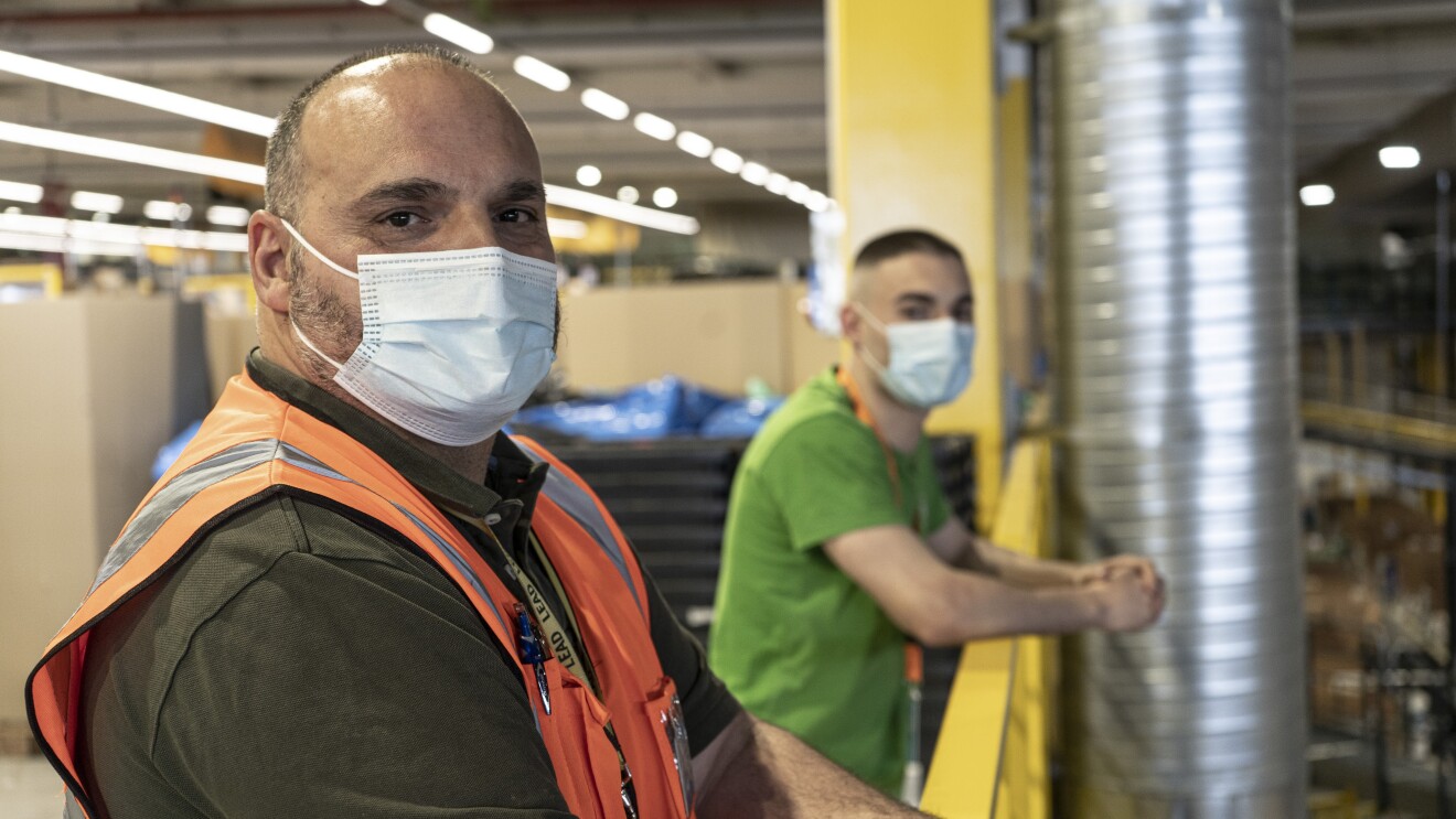 Santiago And´res y Santi Andrés en el centro logístico de Amazon en San Ferando de Henares (Madrid). Desenfocado, de fondo, las estanterías amarillas de un centrlo logístico. En primer plano el padre, Santiago Andrés con pelo muy corto negro y blanco y un poco calvo. Lleva un polo de manga corta de color verde, chaleco naranja y mascarilla. Su hujo en segundo plano con camiseta verde más destacada y el badge con una cinta naranja y mascarilla. Los dos están apoyados a la barandilla amarilla.