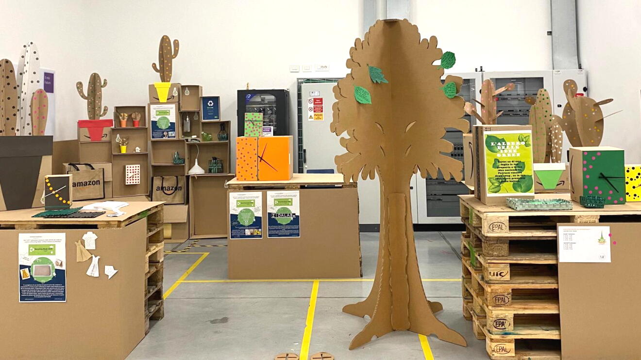All'interno del centro di distribuzione di Vercelli, area realizzata con materiali riciclati tra cui cartone. Al centro c'è la sagoma di un albero, realizzato in cartone, un corner con piante realizzate in cartone e orologi da parete realizzati in cartone. 