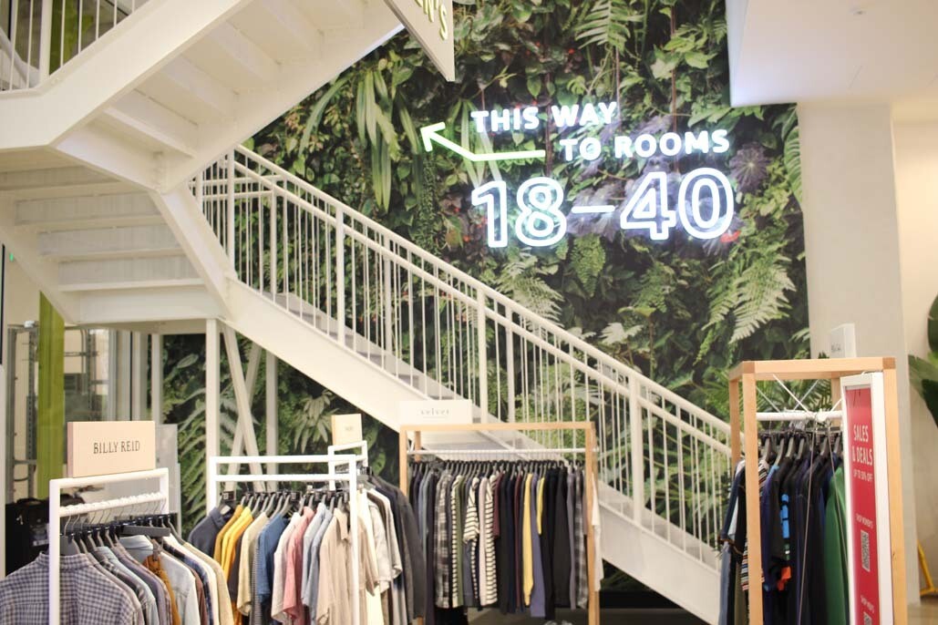 An image of a set of stairs in the Amazon Style store. There is a neon sign on a wall behind the stairs that says "This way to rooms 18-40"