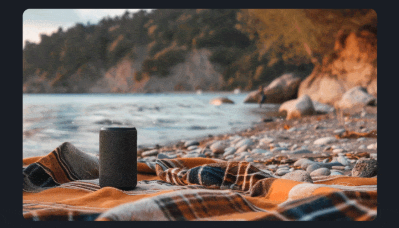 A speaker set on top of a blanket laid out on a the beach. There is w a shoreline in the background.