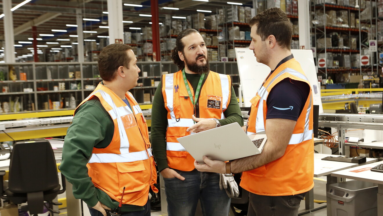 Sergio Amador, lidera el equipo de Health & Safety de los centros de Amazon Operaciones en España. En el centro de la foto con pantalones negros y un chaleco naranja. Rodeado de dos compañeros, el de la derecha lleva un ordenador y un ordenador. De fondo hay meses y estanterías de un centro logístico.