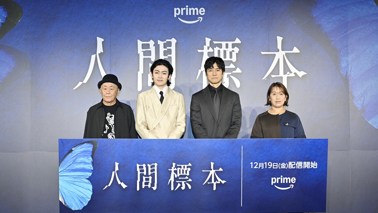 青い壁に「人間標本」の大きな文字。その前に3人の男性とその右側に女性1人が立っている。4人の前には、青い蝶の羽をあしらった青いパネルがあり、白い字で「人間標本」と書かれている。