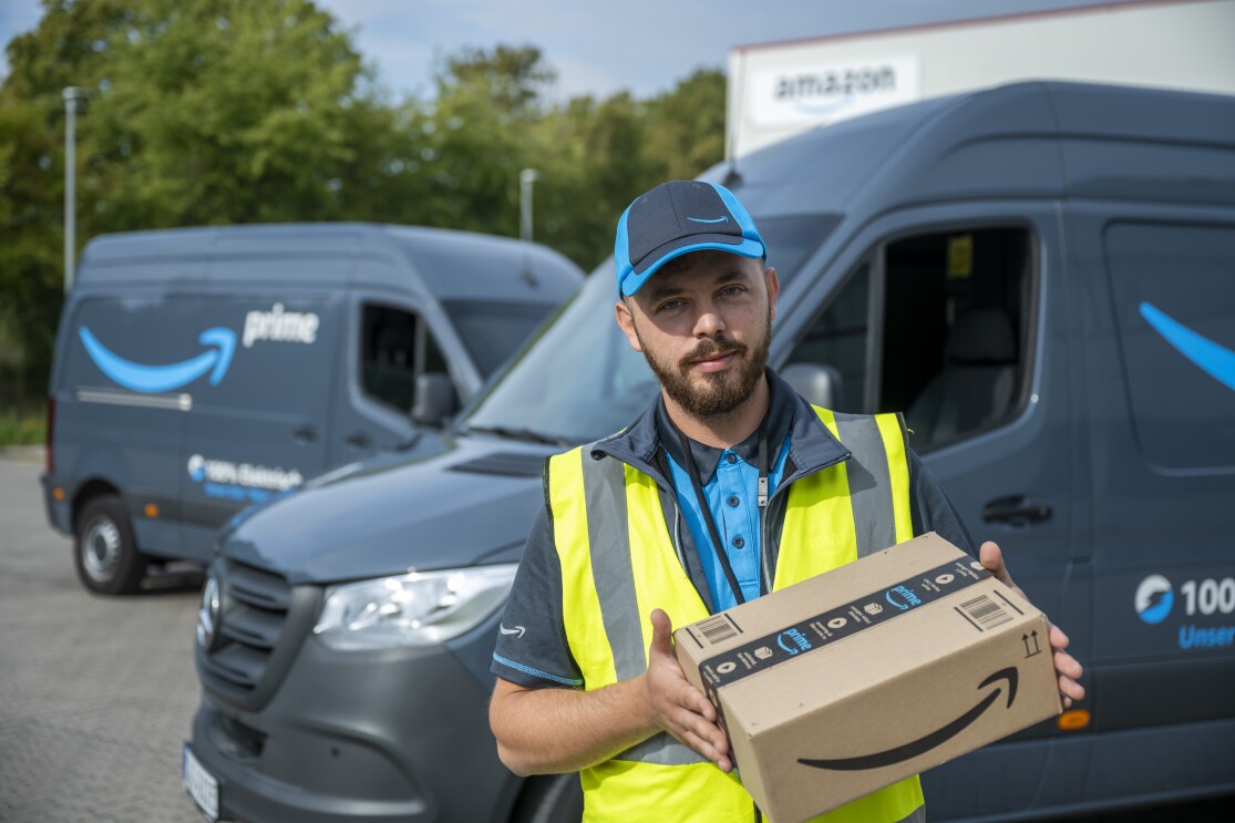 Ein Amazon Lieferfahrer mit einem Paket steht vor einem blauen Lieferfahrzeug.