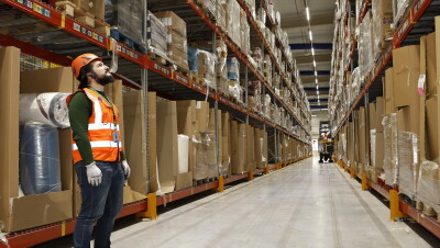 Sergio Amador lidera el equipo de Health & Safety de los centros de Amazon Operaciones en España. Va con casco naranja, chaleco y guantes. Observa las estanterías llenas de cajas. 