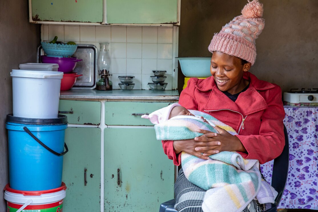 An image of a woman smiling while looking down at a baby she is holding in her arms. The baby is wrapped in a blanket and is wearing a beanie on its head.