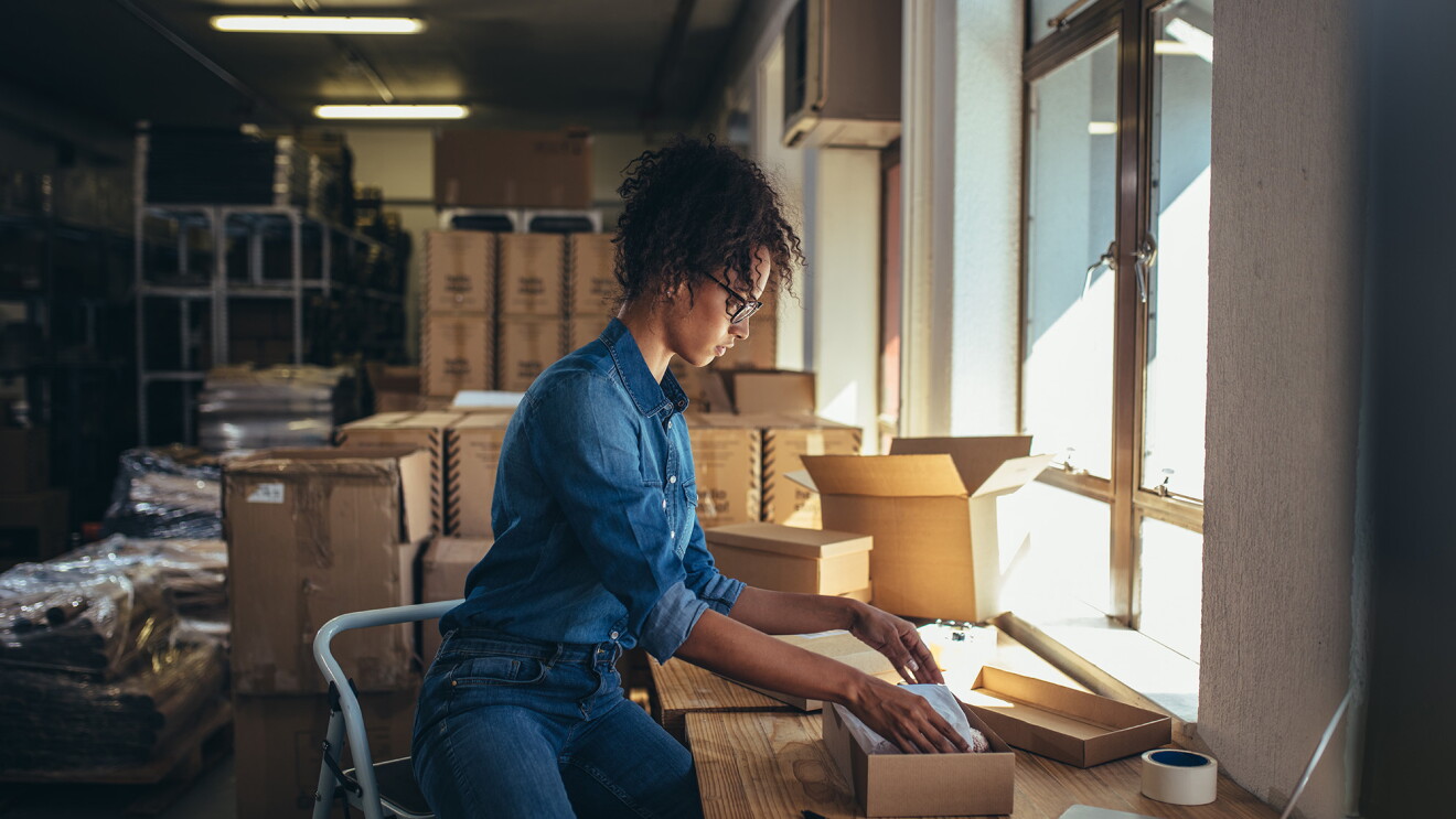 A woman sits on a step stool as she packages a product in a kraft box. 