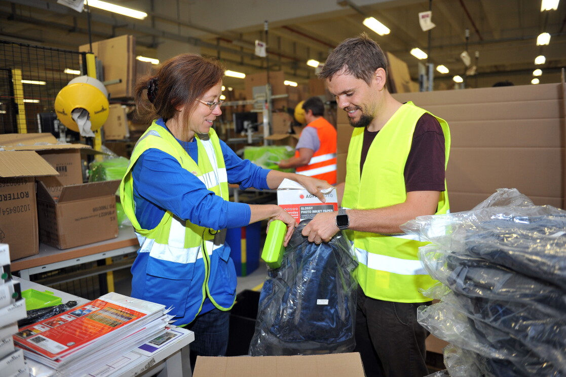 Eine Logistikmitarbeiterin in gelb-blauer Weste packt mit ihrem Kollegen aus der Verwaltung. Er trägt eine gelbe Sicherheitsweste.