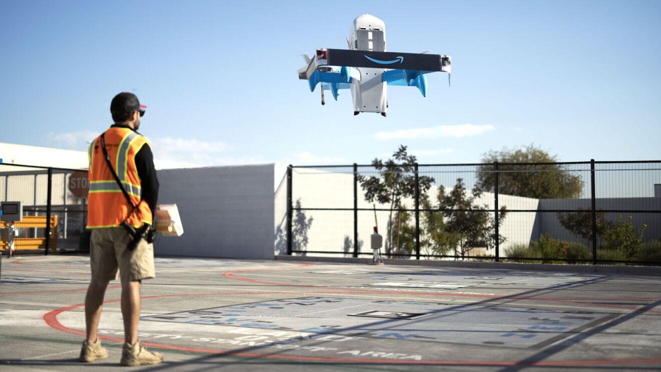 Aerial view of Prime Air drone taking off with safety-vested observer below.