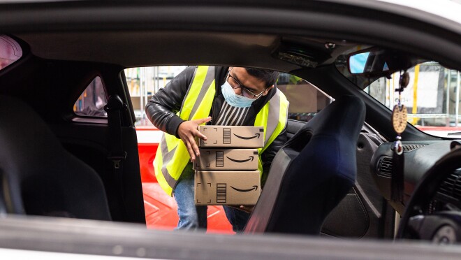 An image of an Amazon delivery driver holding packages while looking into his car. He is wearing a neon safety vest.