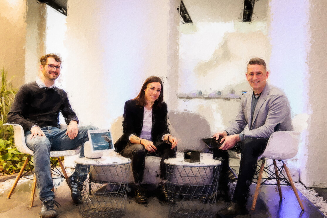 Fotomontaje imitando un cuadro con pinceladas. Equipo de Amazon Lab 126 en España. En la foto, el interior de un espacio de descanso de unas oficinas. Aparecen cuatro personas; tres hombres y una mujer. Están todos sentados. En prímera línea hay dos mesas donde en una hay un dispositivo de Amazon. En el centro hay un sofá de dos plazas con la mujer sentada y un hombre apoyado en el respaldo. En el límite izquierdo y derecho del sofá hay dos sillas blancas con las otras dos personas sentadas.  Descripción de todos las personas que aparecen de izquierda a derecha. El primero lleva unas deportivas, unos jeans, una camisa con un jersey, gafas y el pelo corto y barba de dos días. La mujer va con unas botas, un patalón y una americana negra. Debajo lleva una camiseta blanca. El pelo largo marrón. El segundo hombre lleva botas y pantalón negro, una americana de color gris y un jersey de punto debajo de color azul. El pelo corto y rubio. En la mano él y la mujer están sujetando un dispositivo Amazon. 