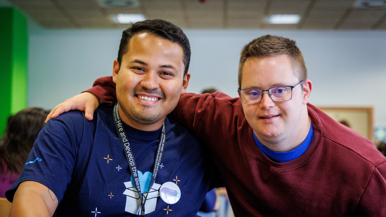 En las instalaciones de Down Madrid en Madrid aparecen 2 personas en primer plano. Un trabajador de Amazon y un participante de la fundación . El trabajador de Amazon van con la misma camiseta azul  y el badge colgado y el de Down Madrid con deportivas con un jersey de color morado. La camiseta del trabajador de Amazon lleva un dibujo que es una caja de Amazon de la que sale un corazón y debajo las palabras Amazon Volunteer. 