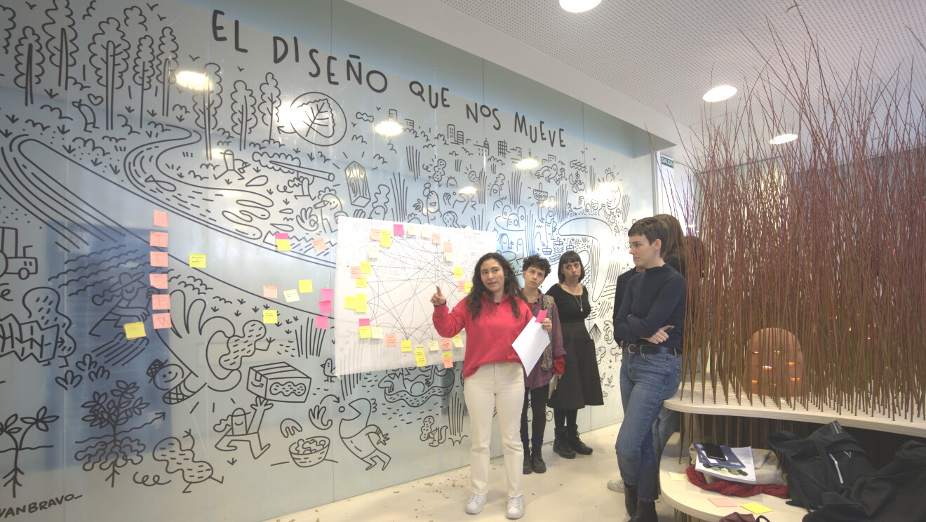 En una pared de cristal con unos dibujos hecho simulando un rotulador debajo del texto El diseño que nos mueve, hay un papel blanco colgado en una pared. El papel tiene papelitos de colores enganchados. No se puede el texto pero todos ellos están conectador por líneas de color negro. Delante del papel hay una mujer con papeles en la mano y dando una explicación delante de tres mujeres más. Esta primera mujer tiene el pelo rizado largo, por el hombro, va con un jersey de color rosa y unos pantalones y unas deportivas de color blanco. Las otras tres mujeres van vestidas de negro. En el suelo hay cáscaras de color marrón, una pared de caña y una mesa de color blanco con un  banco a juego.
