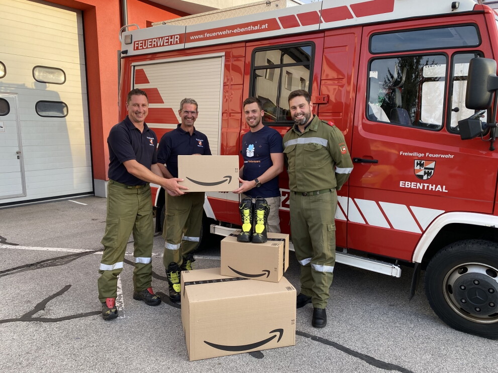 Amazon Mitarbeiter Andreas Widowitz mit Amazon Volunteers T-Shirt übergibt vor einem Feuerwehrfahrzeug Spenden in Amazon Kartons an Kommandant-Stellvertreter Marjian Kropiunik und Kollegen der Freiwilligen Feuerwehr Ebenthal.