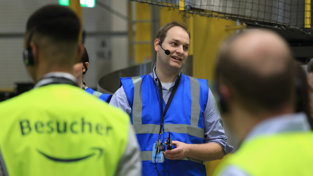 Ein Tourguide in blauer Sicherheitsweste steht vor Besuchern, die eine gelbe Sicherheitsweste tragen. 