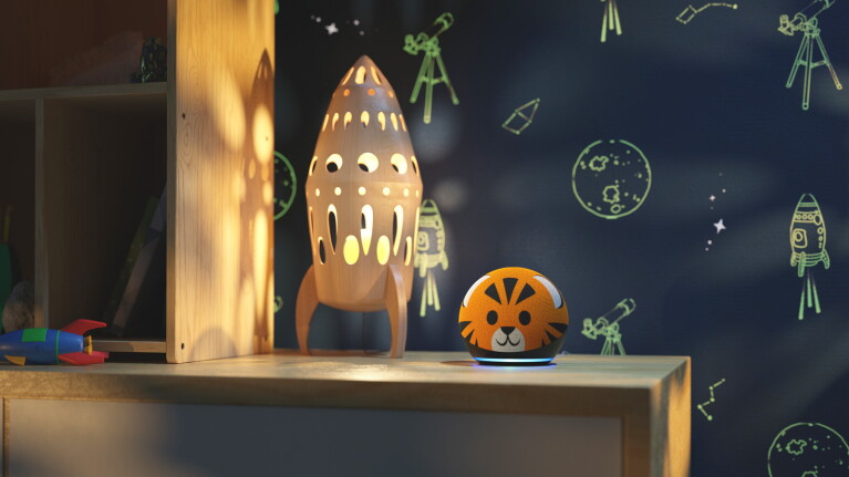 An image of an Echo Kids device next to a rocket shaped lamp sitting on a wooden table. The background is a dark blue wall with green space illustrations including a rocket, a planet and a telescope.