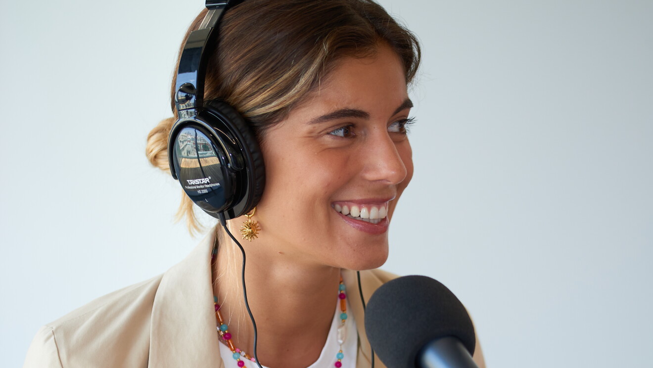SARA BACEIREDO fundadora de It's lava, sentada en una mesa delante de un micrófono y con unos auriculares puestos. Ella está mirando al infinito. Lleva el pelo largo recogido con una cola baja. El pelo castaño con mechas rubias. Una camiseta de punto de color blanco y una maericana beige. Lleva unc ollar de colores y unos pendientes.  