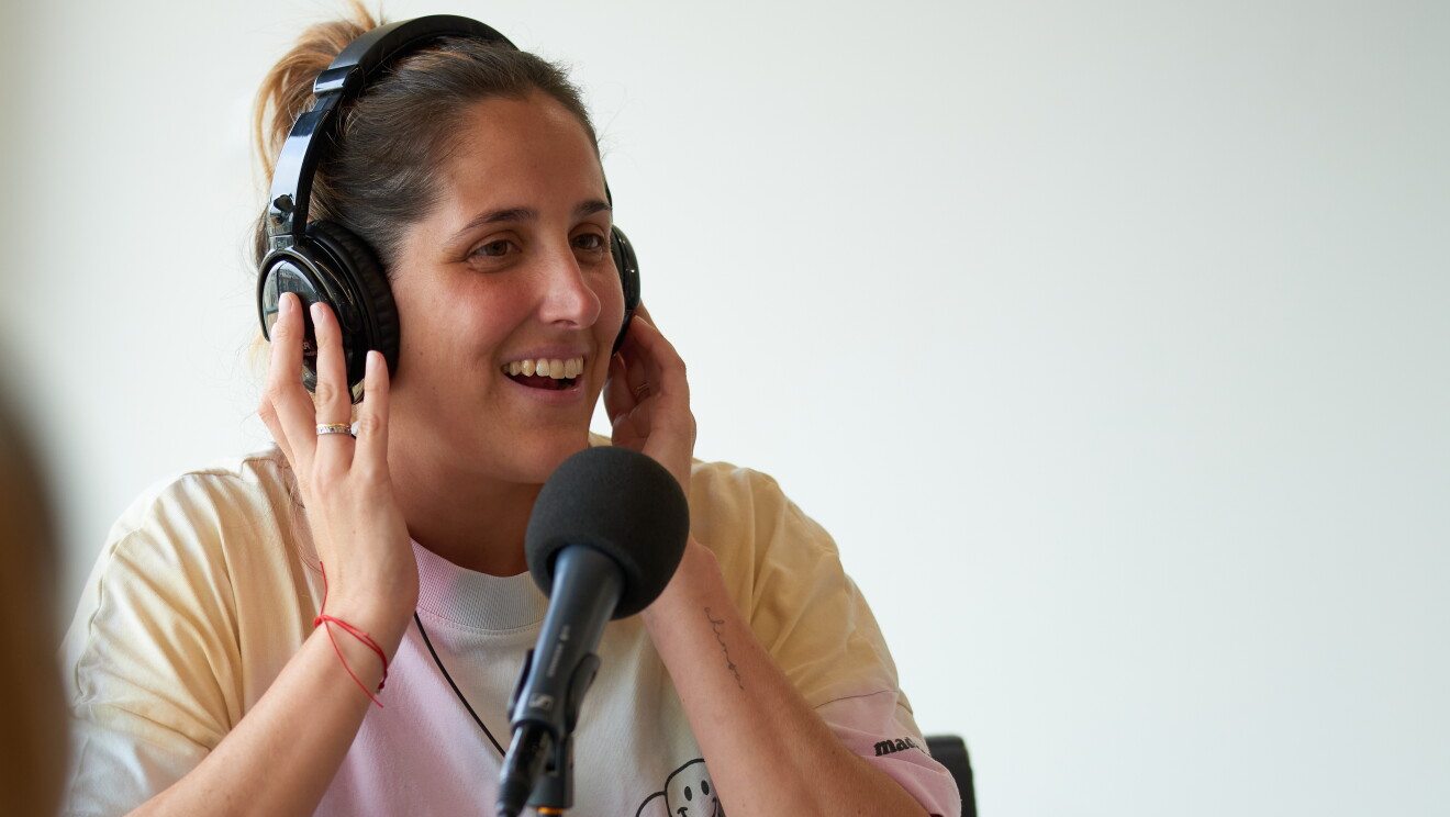 Itziar Aragón, fundadora de Madreamiga. Viste una camiseta blanca con el dibujo de un croassant, el pelo castaño recogido con un moño, un collar y piercings en las orejas. Con las dos manos en los auriculares y delante de un micrófono. Está sonriendo, lleva un anillo en la manoderecha con una pulsera roja. 