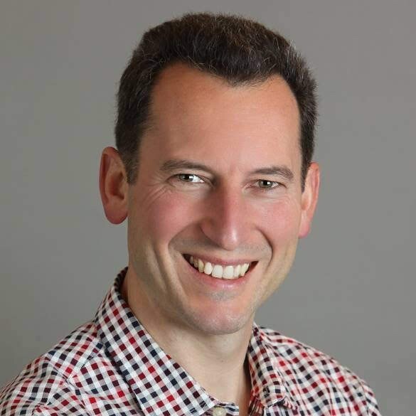 Alex Smola, machine learning director for Amazon Web Services’ Deep Engine group wears a blue striped hooded sweatshirt over a black tee shirt, and a cross-body bag. He stands outside near a rock formation.