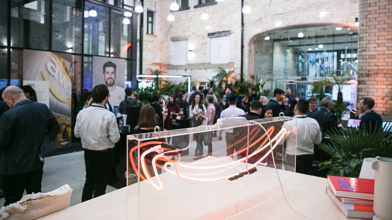 A photo of Amazon employees standing and chatting at the Manchester Corporate Office opening. On a table, there is a large Amazon smile logo. 