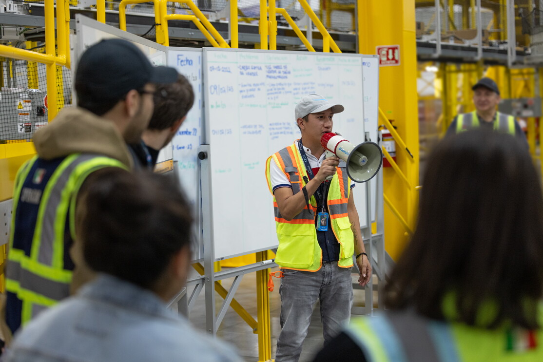 En el centro logístico Mex6 se ven difuminados y de espalda a cinco trabajadores con sus chalecos de color amarillo. En el centro de la foto un trabajador de Amazon con un megáfono hablando. Va vestido con el chaleco de seguridad, una gorra de color clarito, un polo blanco, rojo y azul, y unos jeans grises. Está hablando a sus compañeros. Detrás de él hay tres pizarras escritas con rotulador azul y verde. De fondo están las estanterías donde se almacenan los productos. La estructura es de metal amarillo.