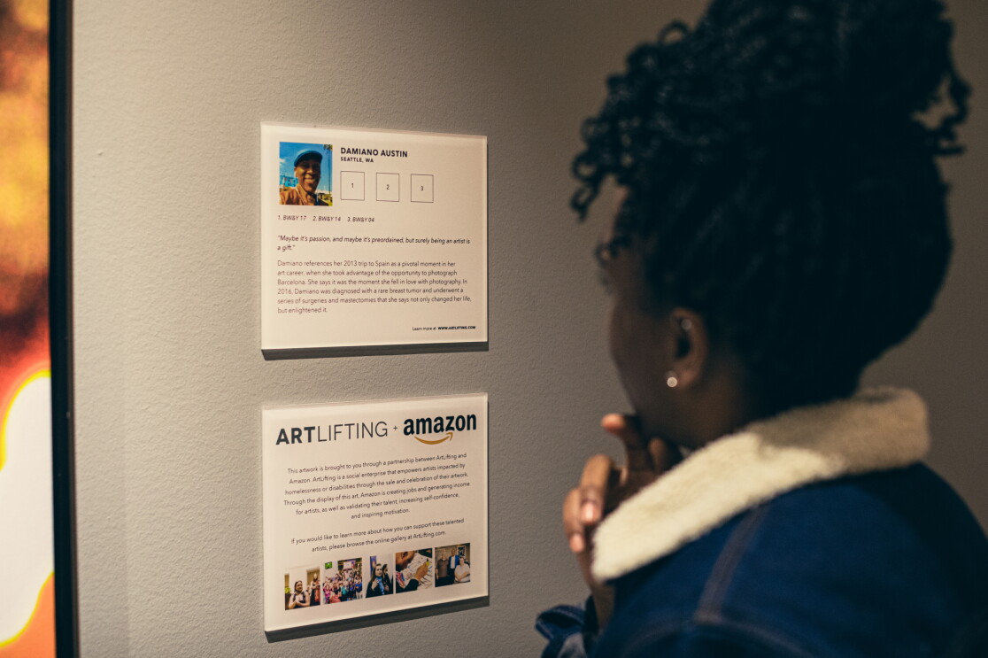 A woman in a blue jacket looks at an artist information card at a gallery. 