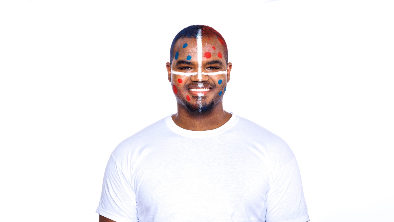 Para el Día de la diversida Amazon ha preparado una acción con asociados de diferentes países que llevan la bandera pitnada en su cara. En la foto, Ángel con camiseta blanca, mirando sonriendo a cámara con la bandera de su país puntada en la cara. Tiene el pelo muy corto y perilla. 