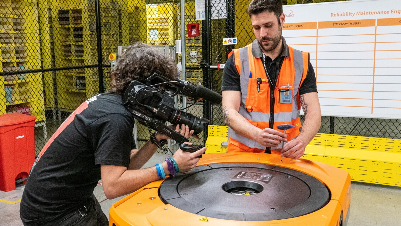 Raúl Cruz, técnico senior de mantenimiento en robótica, en un momento de la grabación mientras manipula un robot. En primer plano está Raúl, con el pelo negro y barba, vestido con una camiseta negra y el chaleco naraja. En la mano tiene una herramienta para abrir un robot. A su lado derecha aparece el cámara, vestido de negro grabando la reparación. 