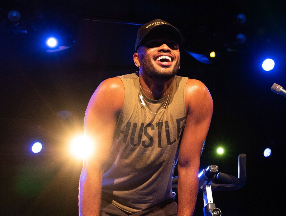 An image of a man smiling while looking out at a group of people. There is a stationary bike behind him and stage lighting.