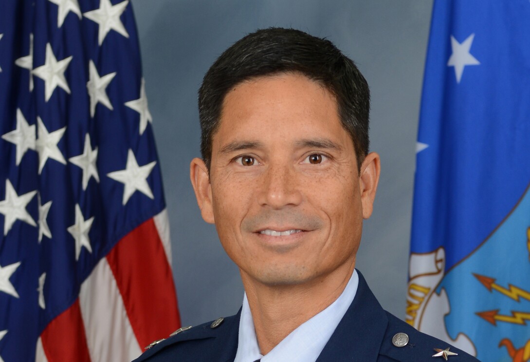 A headshot of a man in a military uniform, smiling for a photo in front of the American flag to his left and another flag to his right.