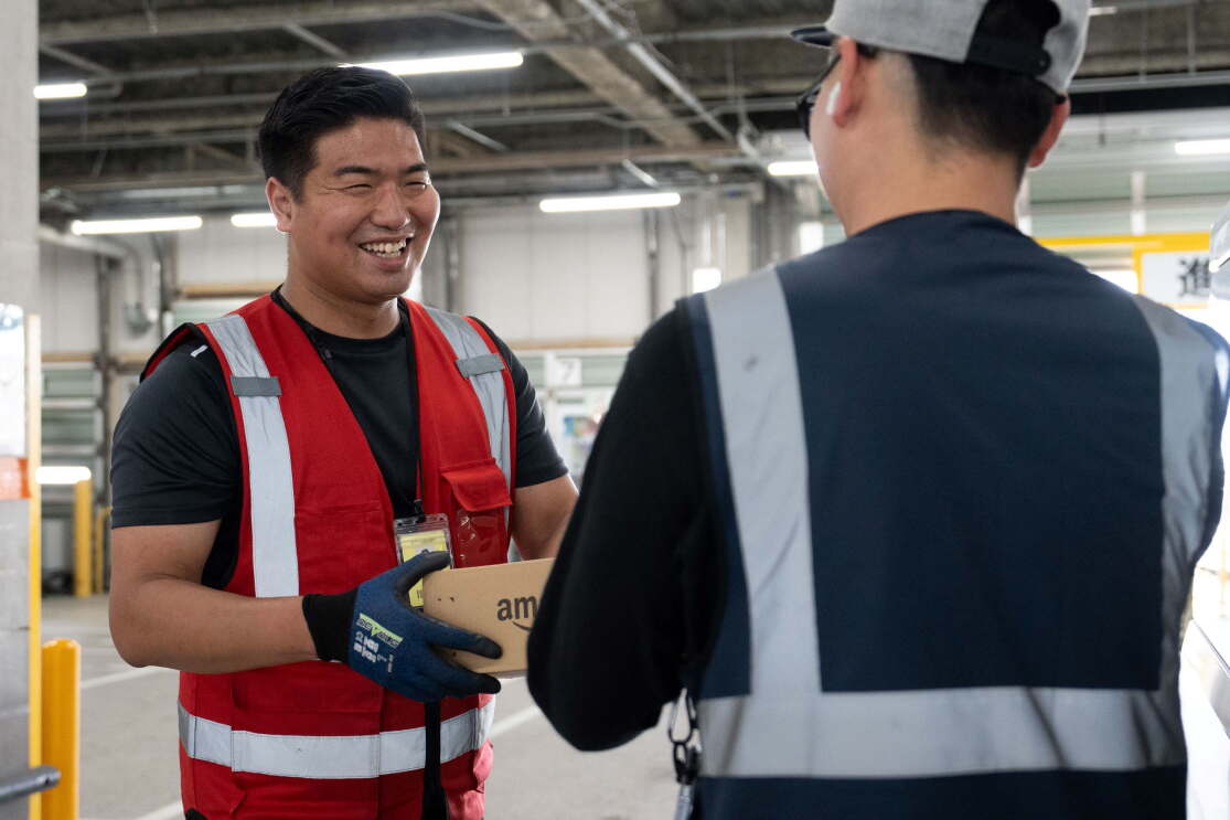 広い空間で赤い安全ベストを着ている男性が青い安全ベストを着ている男性にAmazonの箱を渡している。