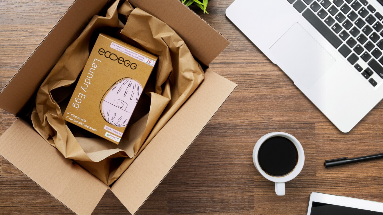 Ein Laundy Egg im Versandkarton von Amazon mit Recycling-Verpackungspapier anstatt mit Plastik Luftpolsterkissen. Daneben liegen ein Laptop, ein Stift, ein Tablet und eine gefüllte Kaffeetasse.