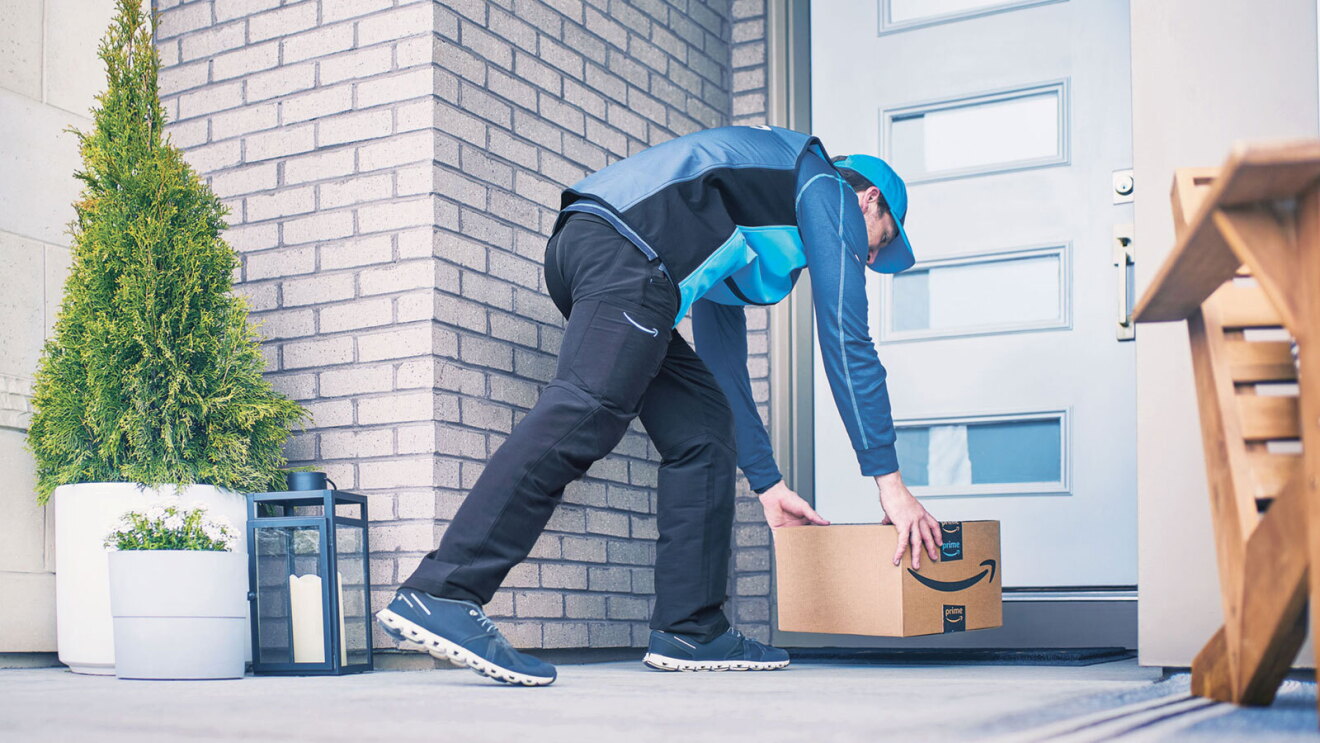 A delivery driver for Amazon bends down to deliver a package on a front porch.