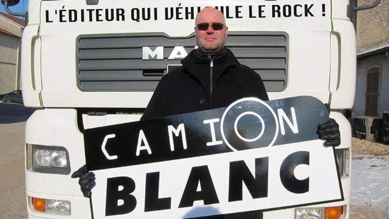 Un homme portant des lunettes de soleil se tient devant un camion blanc en tenant dans ses mains la pancarte de la maison d'édition Camion Blanc