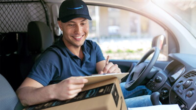 Un repartido de Amazon en una furgoneta. Está sentado en el asiento del conductor. Lleva un polo azul y una gorra de Amazon con la sonrisa en azul. Se está apoyando sobre una caja para escribir en una hora.