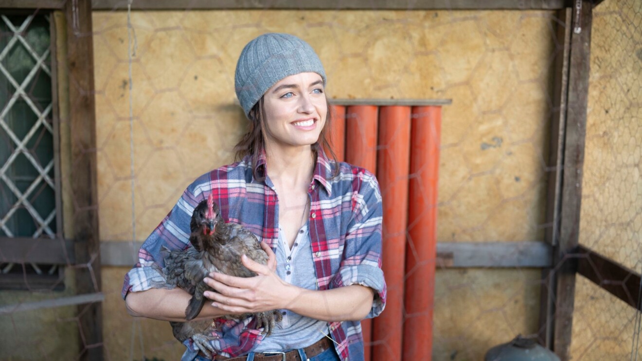 A woman wearing a beanie and holding a chicken in a farm setting