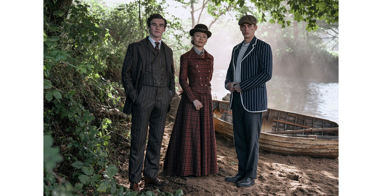 水辺の砂地に立つ、歴史的な服装をした3人の人物。背後の水面に光がさし、ボートが砂地に置かれている。